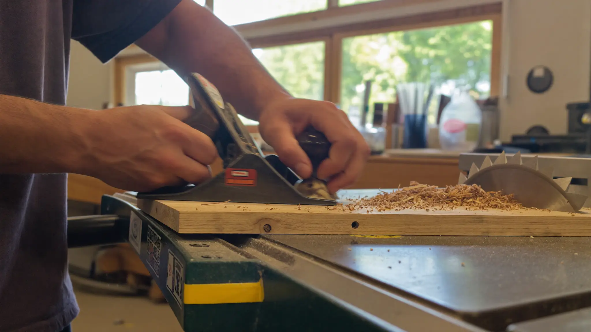 Carpenter's hands guiding a hand plane along a wooden board on a sawhorse, wood shavings curling off the blade