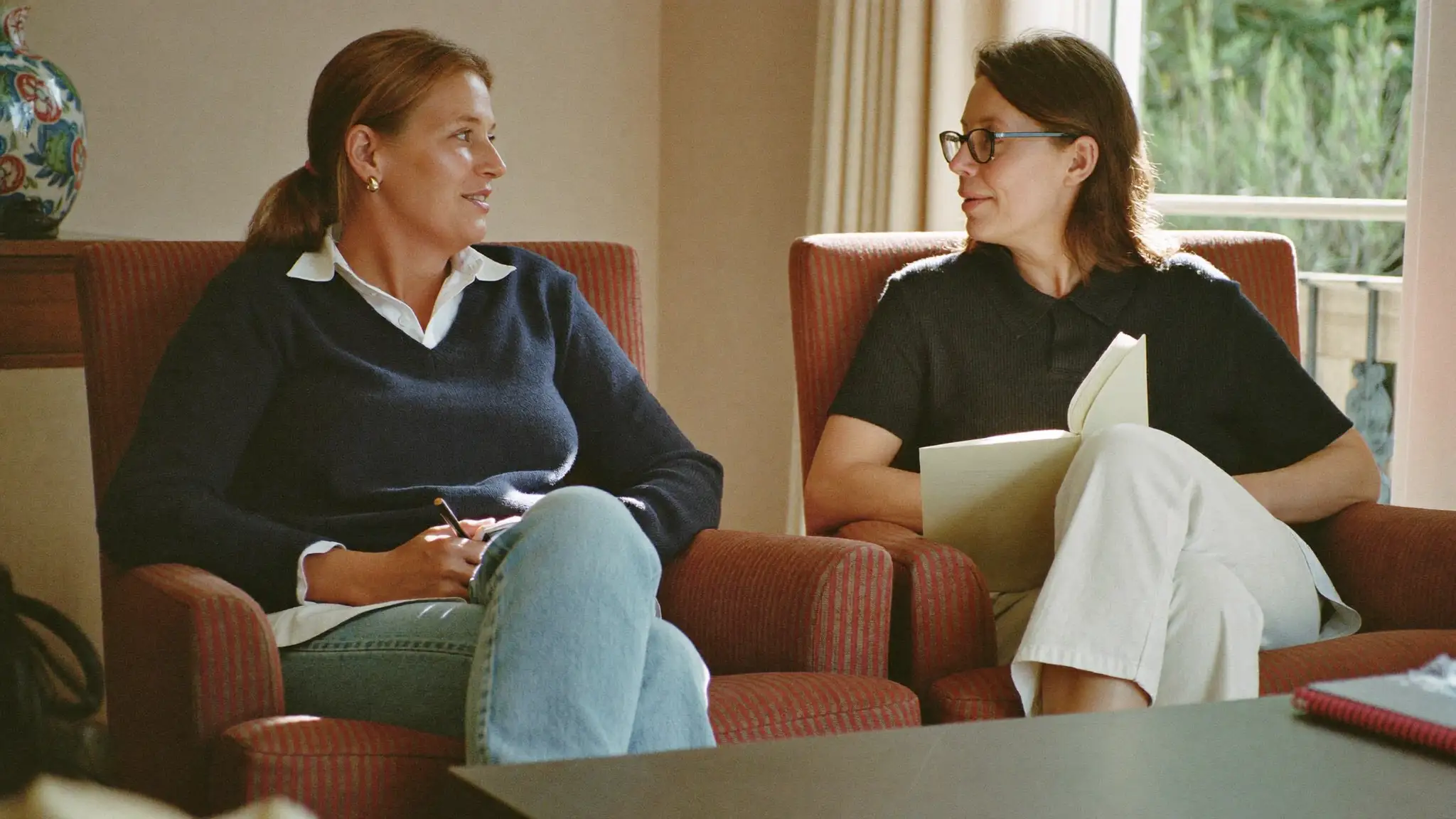 Two women in conversation while seated in armchairs in a sunlit living room