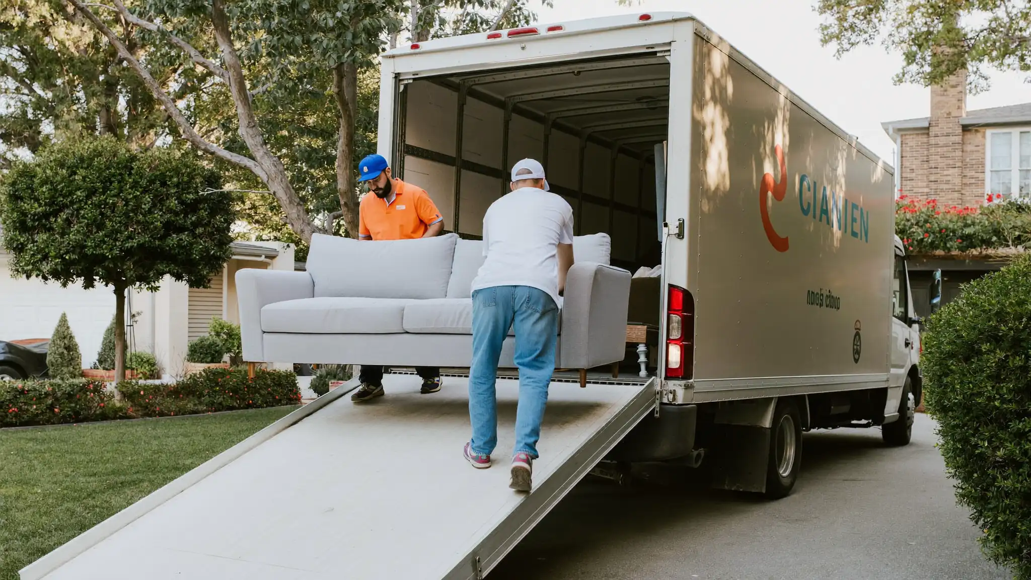 Two movers carrying a wrapped sofa down a ramp into a moving truck parked in a driveway