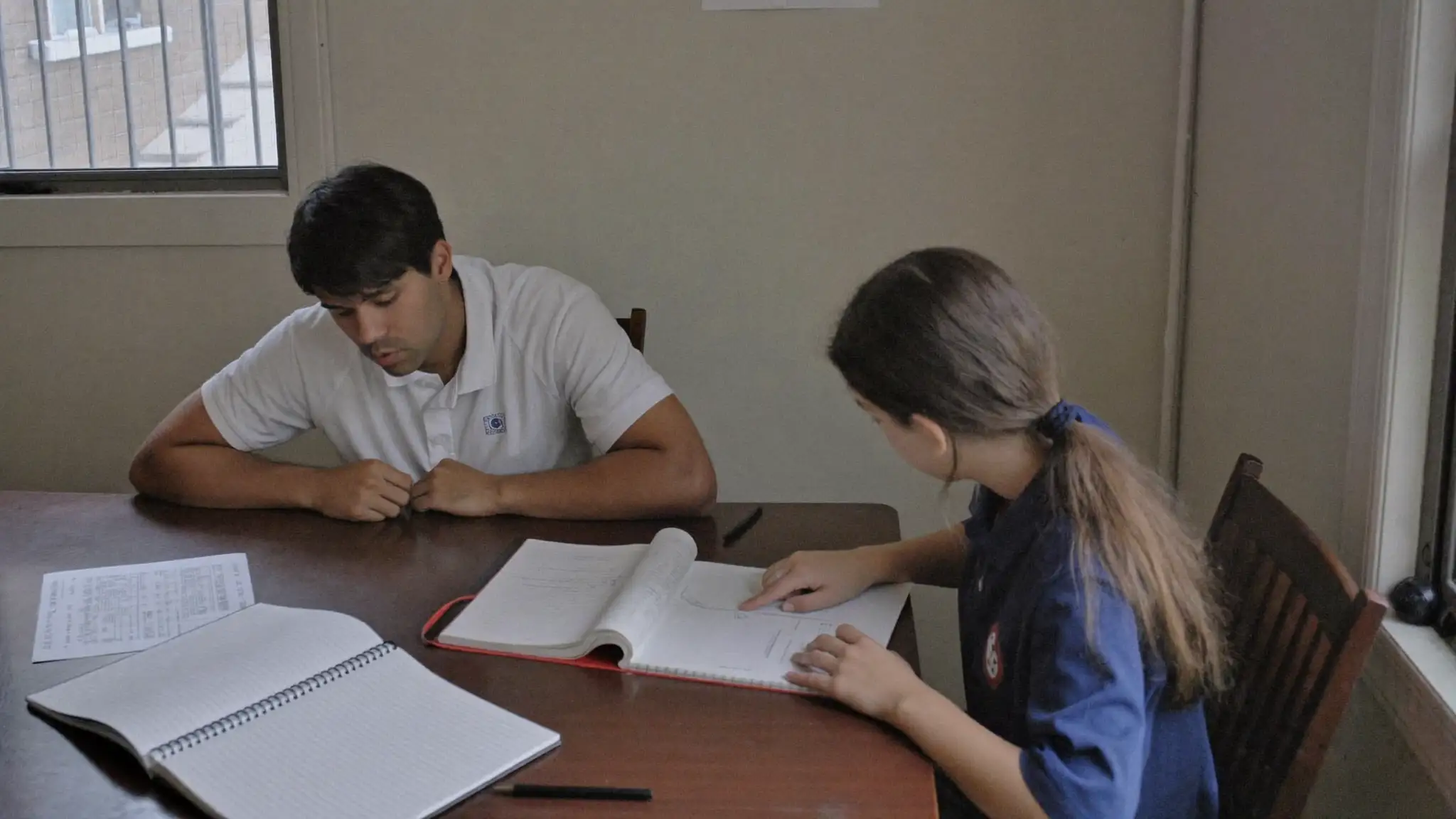 Tutor sitting beside a student at a table, pointing to an open notebook while helping with homework