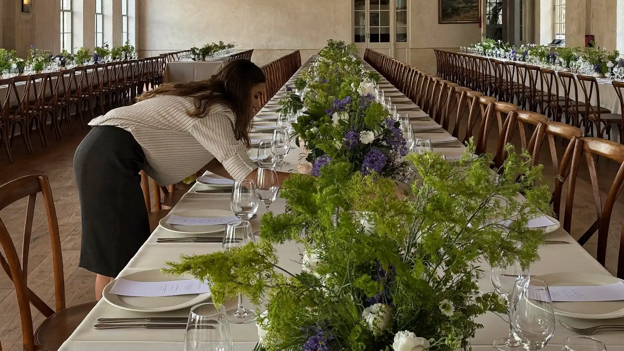 Event planner leaning over a banquet table arranging a floral centerpiece in a sunlit event hall